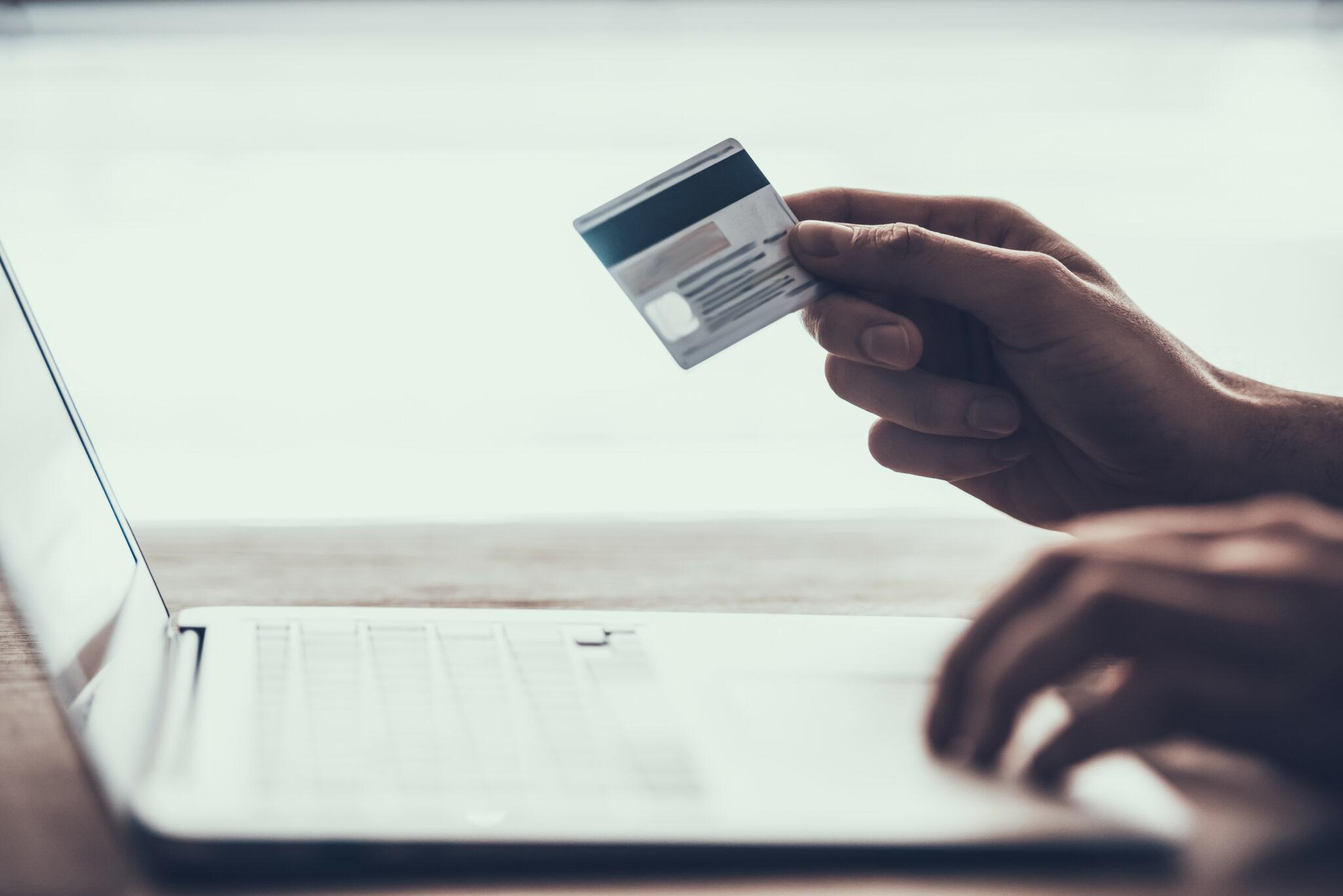 A close-up of a hand holding a credit card in front of a laptop, suggesting online shopping, digital payments, or internet banking, set against a softly lit, minimalist background.