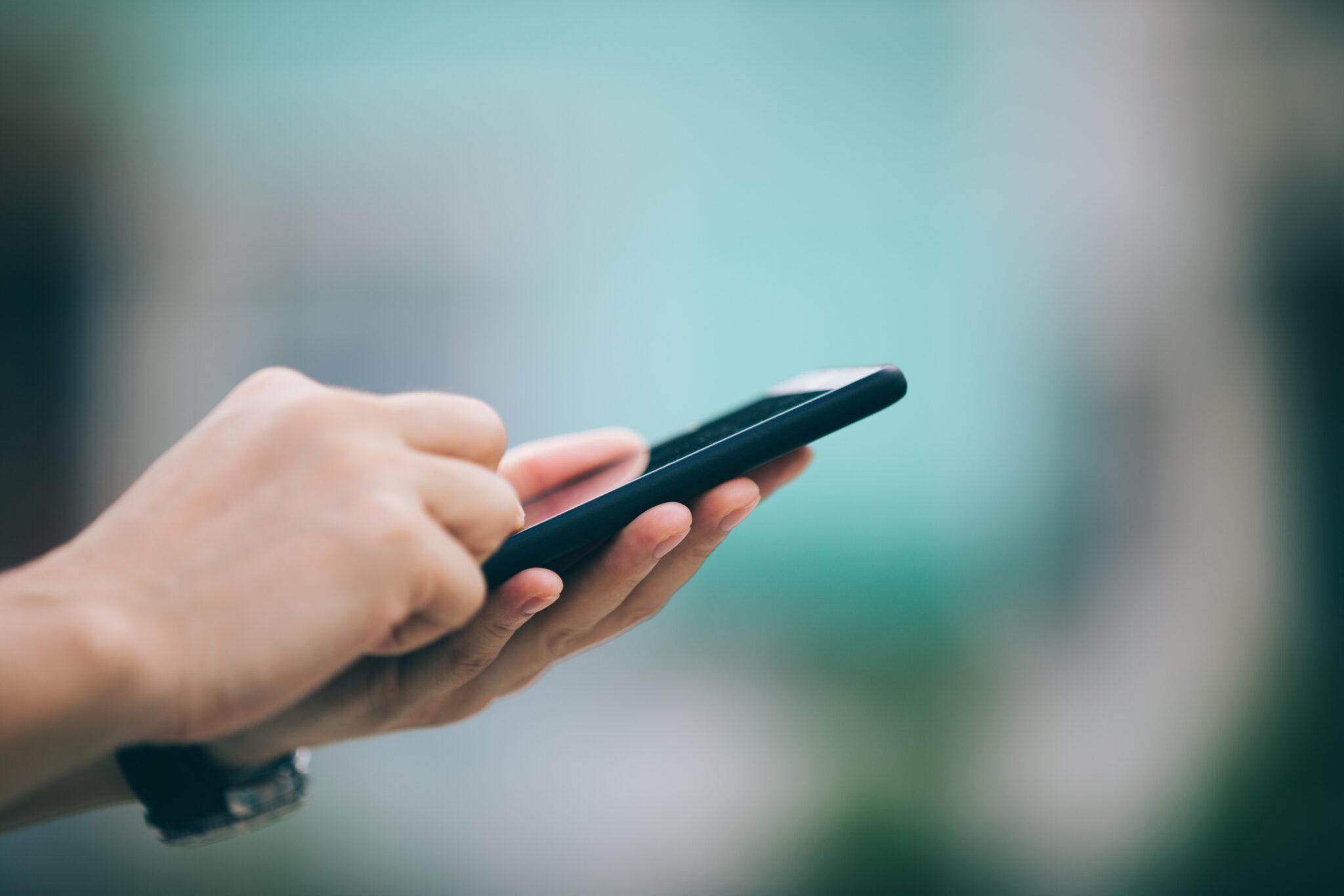 A close-up of hands holding and interacting with a smartphone, set against a blurred, softly colored background, suggesting mobile communication or digital engagement on the go.