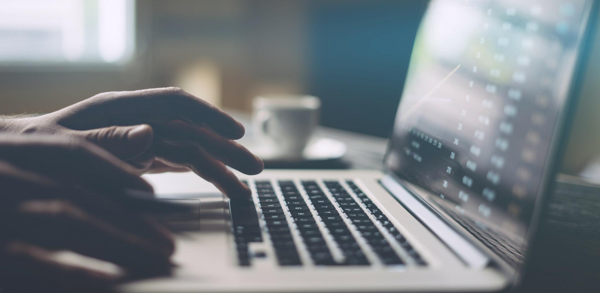 A close-up of hands typing on a laptop keyboard, with a blurred screen displaying data and graphs. A soft-focus coffee cup sits in the background, evoking a calm and focused workspace atmosphere.