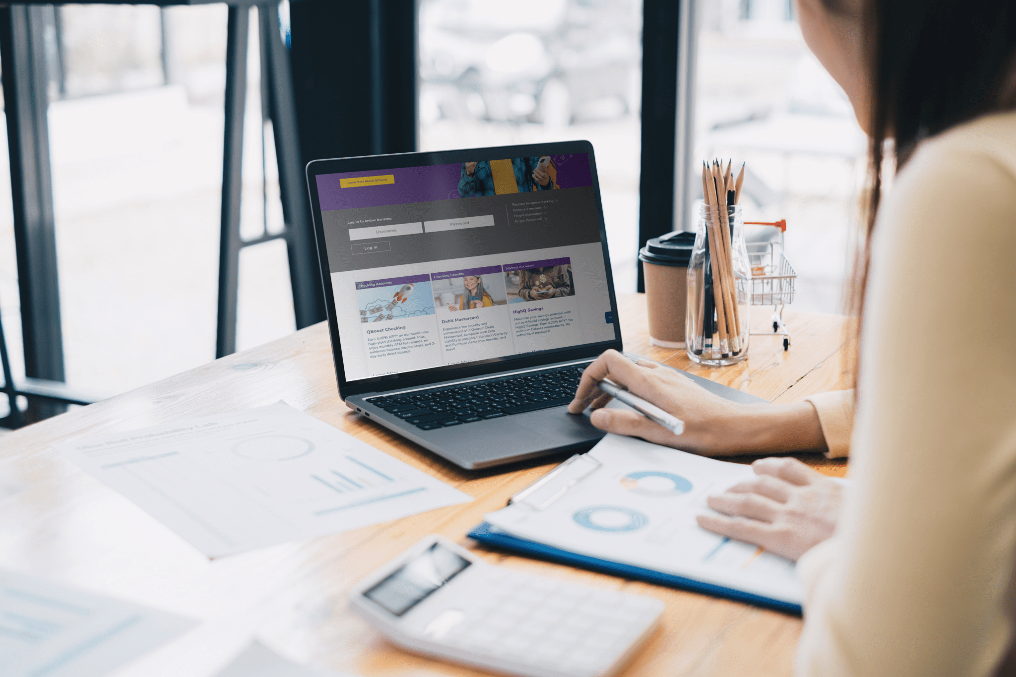 A person working on a laptop at a desk, reviewing a website interface featuring login options and informational sections. The workspace includes documents with charts, a pen, a calculator, a coffee cup, and a jar of pencils, set in a bright, open office or café environment.