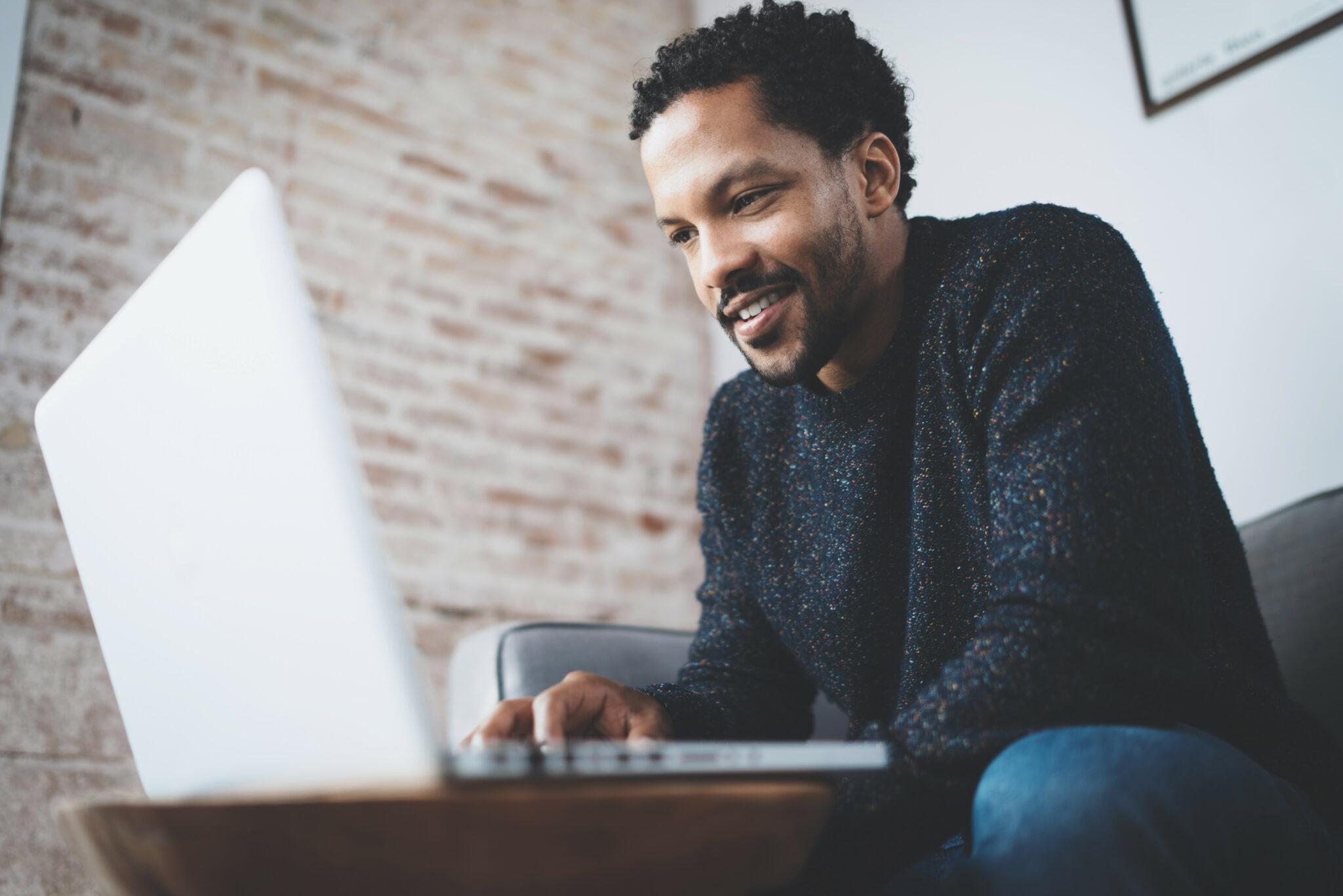 A smiling man in a cozy sweater working on a laptop in a modern, relaxed setting with an exposed brick wall in the background, creating a warm and professional vibe.