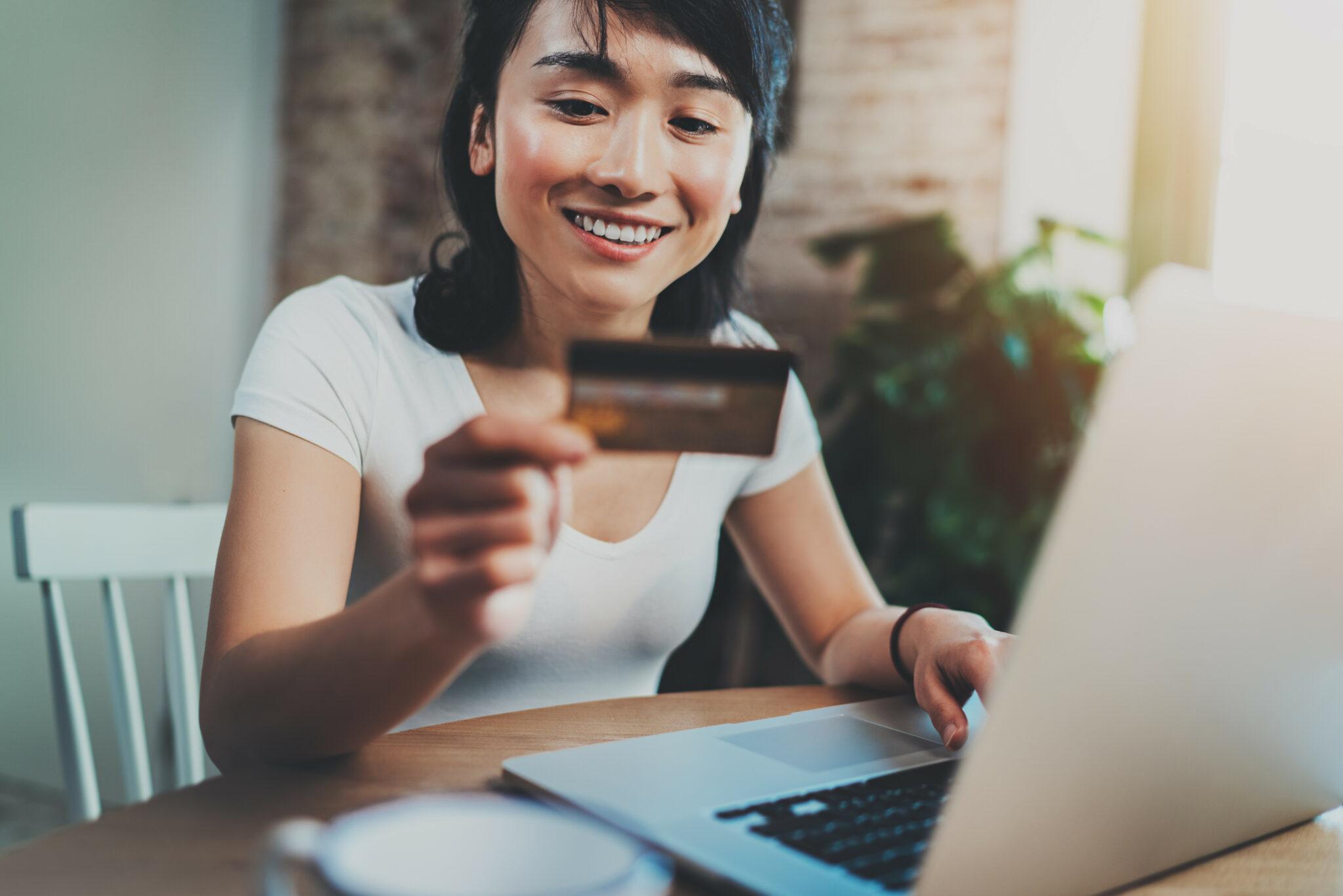 A smiling woman holding a credit card while using a laptop, seated in a cozy and naturally lit space with a warm and inviting atmosphere. The scene suggests online shopping, banking, or a digital transaction.