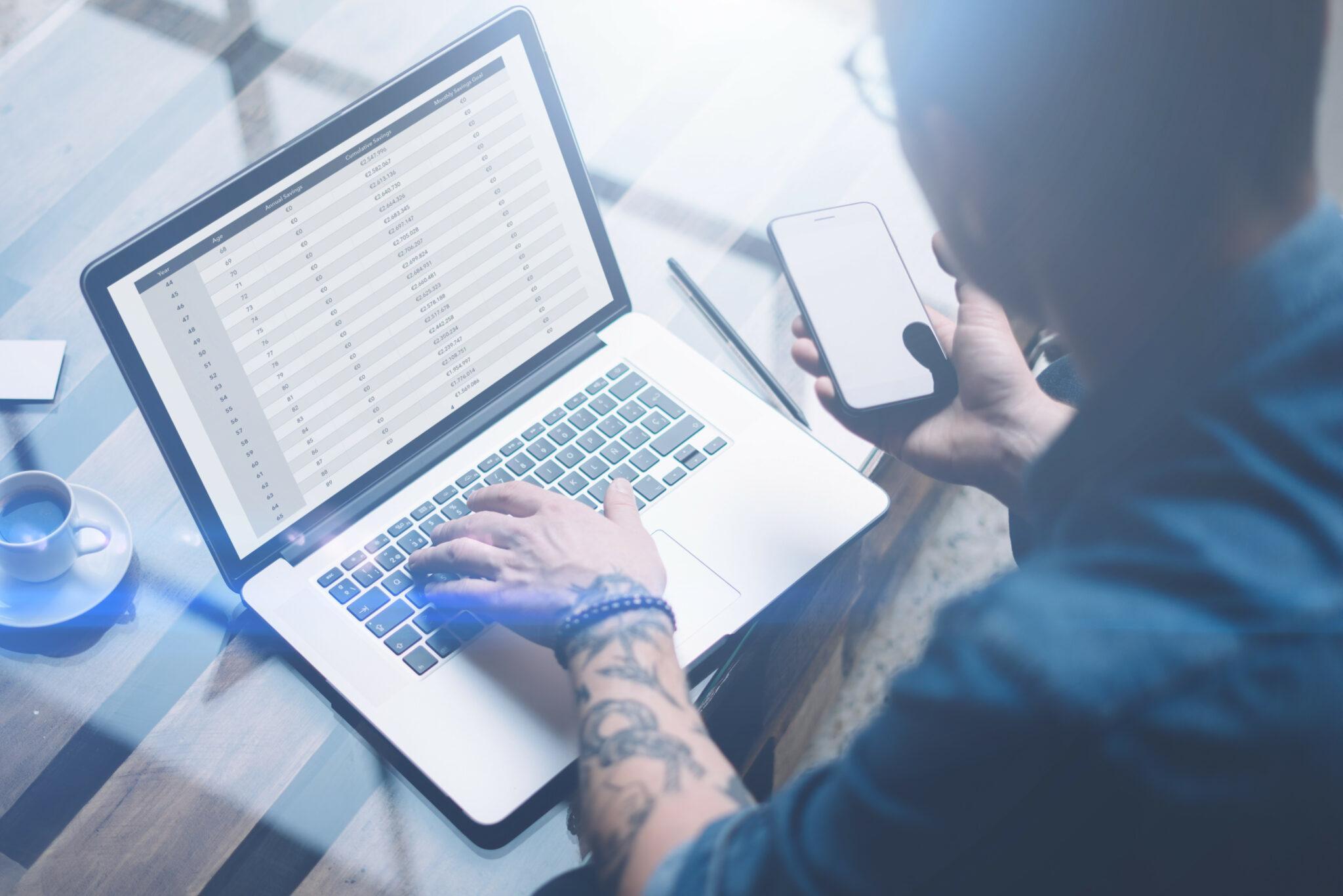 A person working at a laptop displaying a spreadsheet, holding a smartphone in their other hand. The setting is bright and modern, with a cup of coffee and a pen on the table, suggesting a productive workspace.