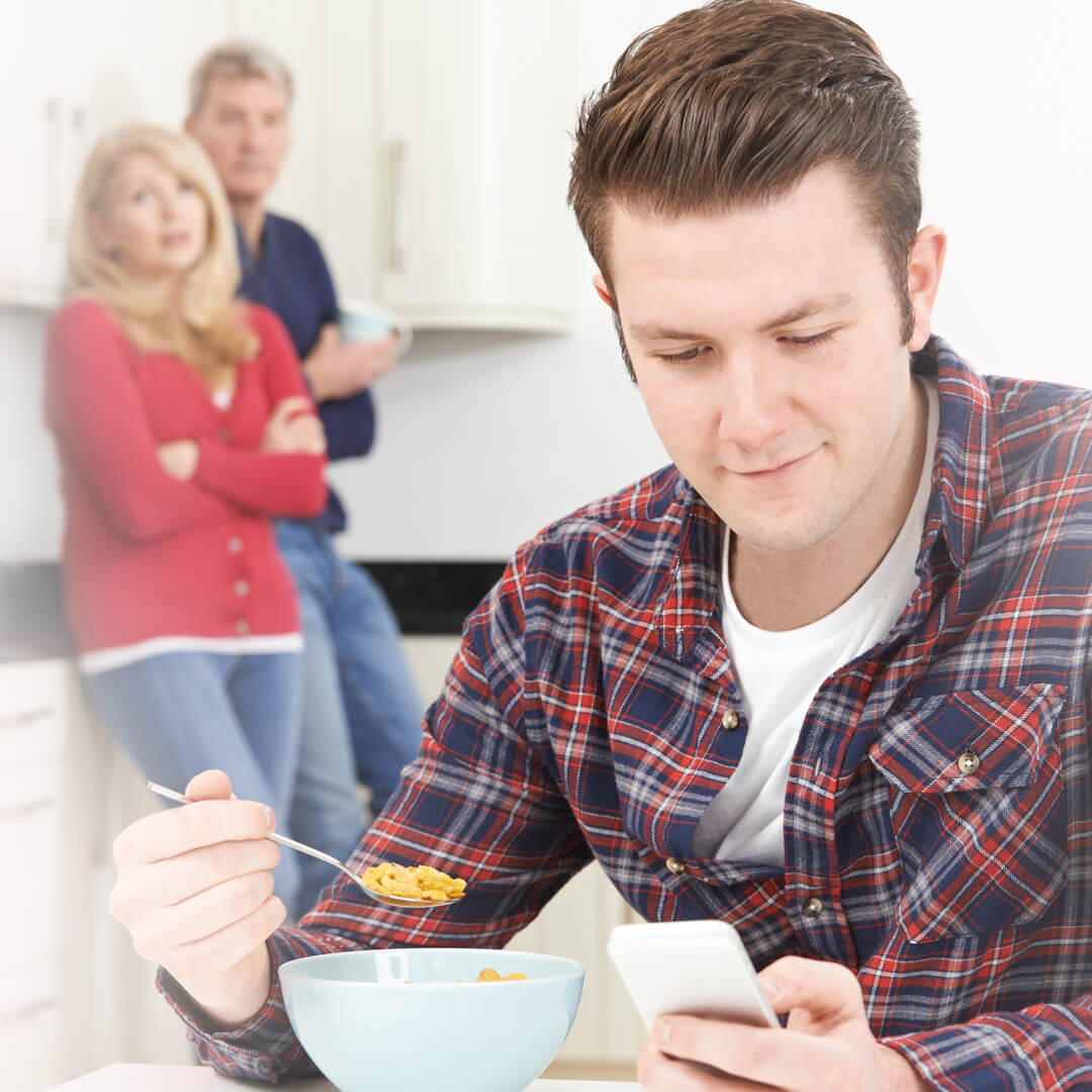 Grown man eating cereal at parents' kitchen table, parents looking at him with concern.