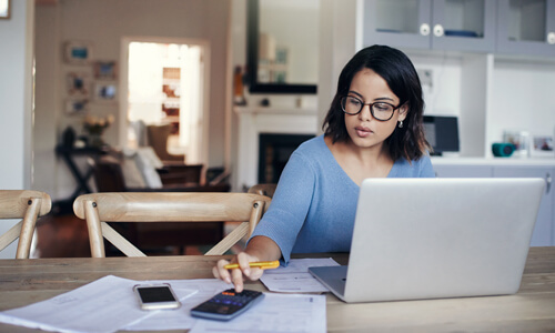 Woman sits at her desk with laptop and calculator, working on her budget.