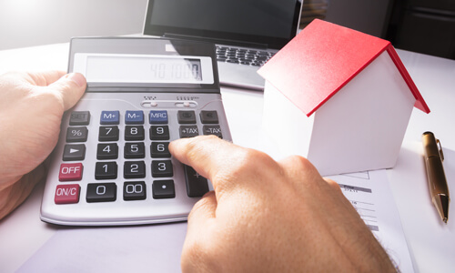 Closeup of hands punching numbers into calculator, wooden house to side.