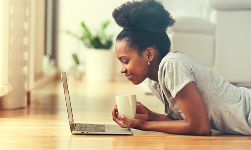 Young woman entrepreneur lying on her stomach on floor with a cup of coffee, looking at her laptop.