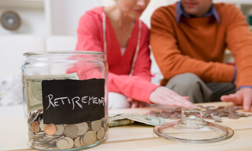 Closeup of change jar labeled "Retirement." Couple in back looking over more coins.