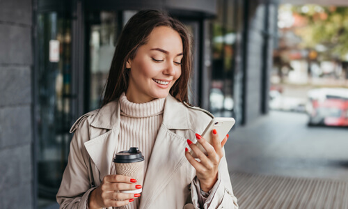 Smiling woman holding coffee and shopping, smiling at her mobile phone, checking her available balance.