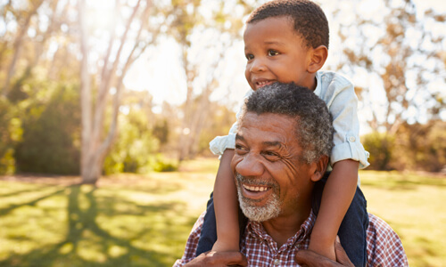 Grandfather with grandchild on shoulders, both smiling outdoors.