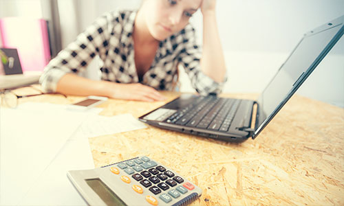Stressed woman looking at laptop and calculator after incurring unexpected household expenses.